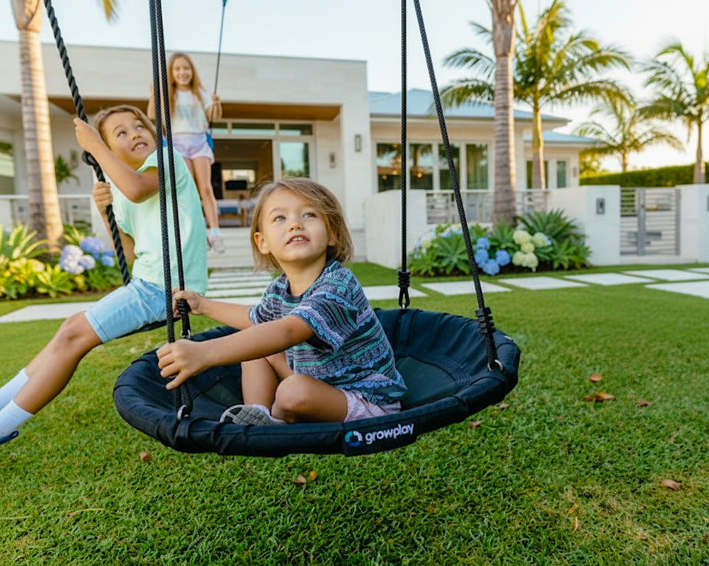 Children playing on a swing set in a backyard with a house and palm trees in the background.