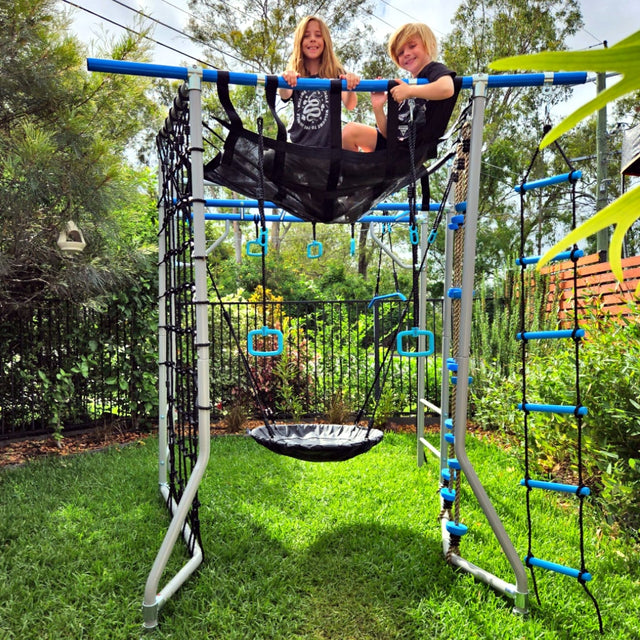 Two kids sitting on the Ninja Net on top of a blue Compact frame in a leafy backyard