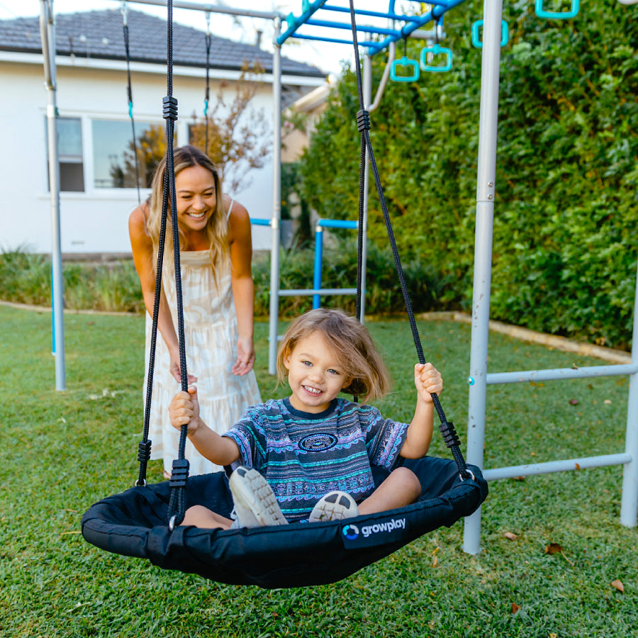 Child in a swing set with a woman standing behind them in a backyard setting.